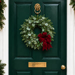 A green eucalyptus wreath with a large red velvet bow, hanging on a dark green front door with a gold lion knocker and brass mail slot.