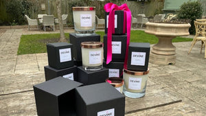 A wooden looking table has several candles stacked onto of each other in a display. The candles are in sleek black boxes with rose gold lids, and one is tied with a pink ribbon. Inspired by Adam's favorite flowers, the setup glows against a blurred outdoor backdrop.