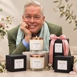 A person smiles while leaning on a table with New Devine Candles by Cookham Handcrafted Candles. The candles are in sleek black boxes with rose gold lids, and one is tied with a pink ribbon. Inspired by Adam's favorite flowers, the setup glows against a blurred outdoor backdrop.