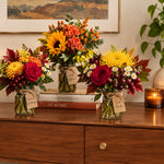 Four autumn floral arrangements in clear glass jars with twine tags, displayed on a wooden console table in a warmly lit room. The bouquets include sunflowers, roses, daisies, burgundy grapes, and fall leaves.